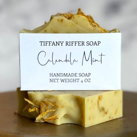 Two yellow calendula mint soap bars sitting in front of white blurred background. The widest surface of one bar faces the viewer to show the clean white label and branding for Tiffany Riffer Soap. This handmade vegan soap contains calendula petals inside and on top of the soap bar for a natural and gentile exfoliation. Each bar weighs approximately 4.0 ounces, and are approximately 3.5 inches long, 1 inch wide, and 2.5 inches tall.